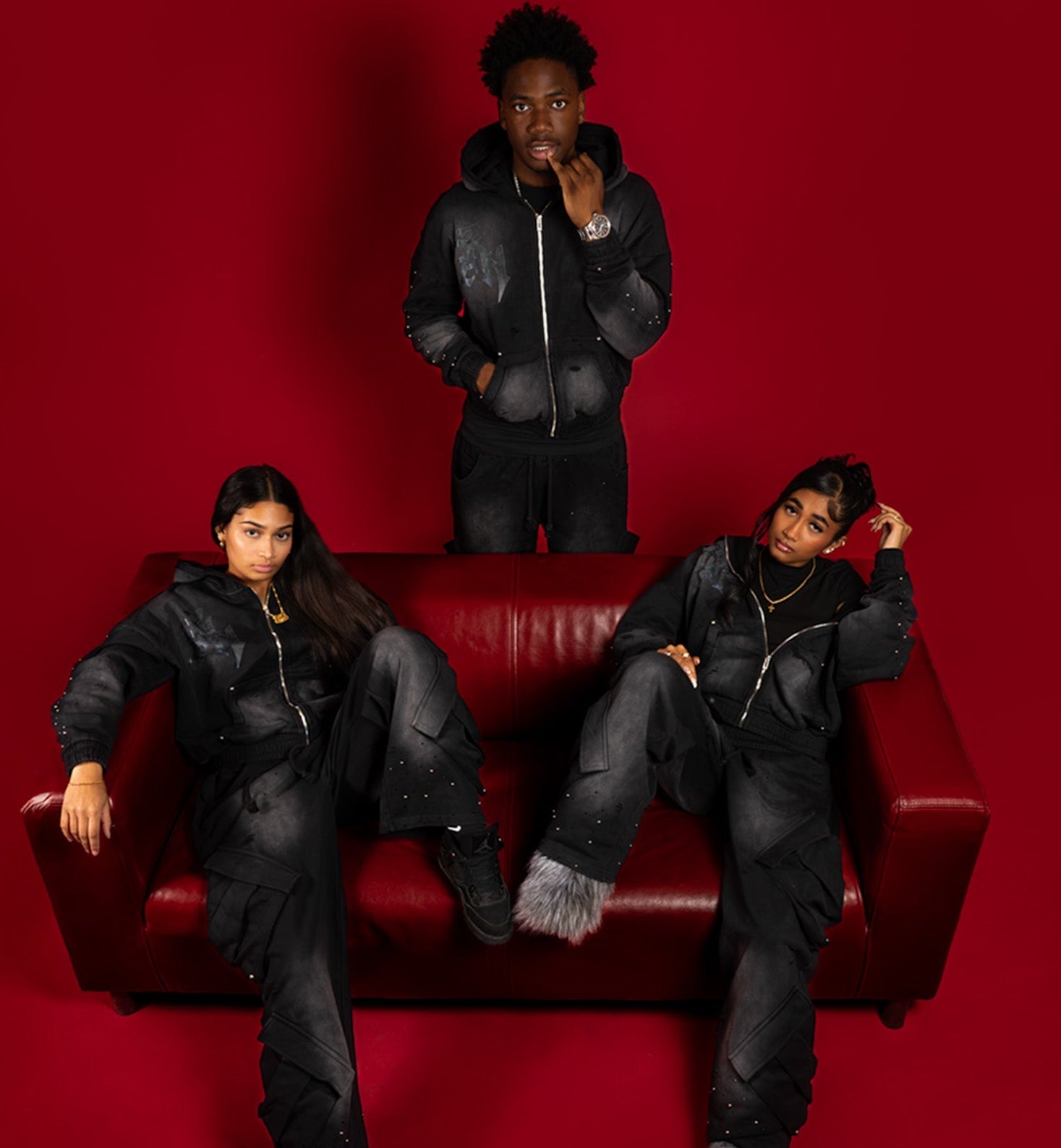 Three people in black outfits sitting on a red couch against a red background